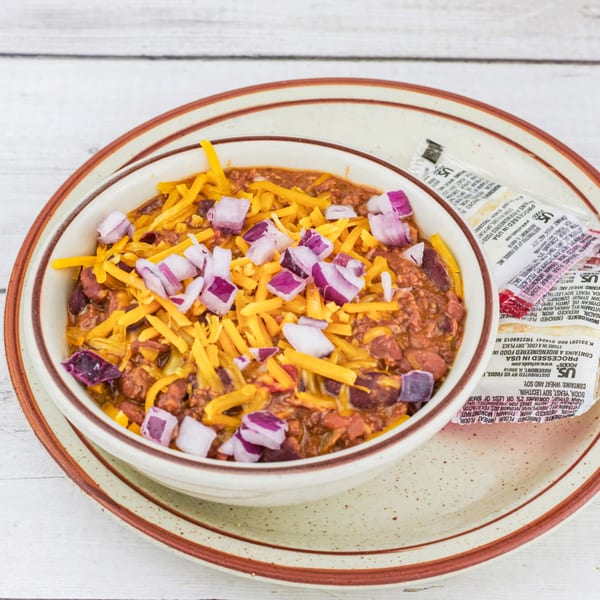 A bowl of chili topped with shredded cheddar cheese and diced red onions, with condiment packets on the side.