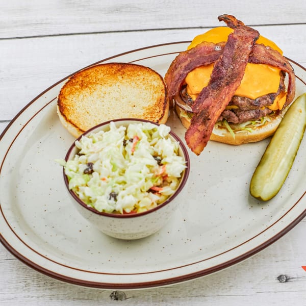 A bacon cheeseburger with a side of coleslaw and a pickle spear on a white plate.